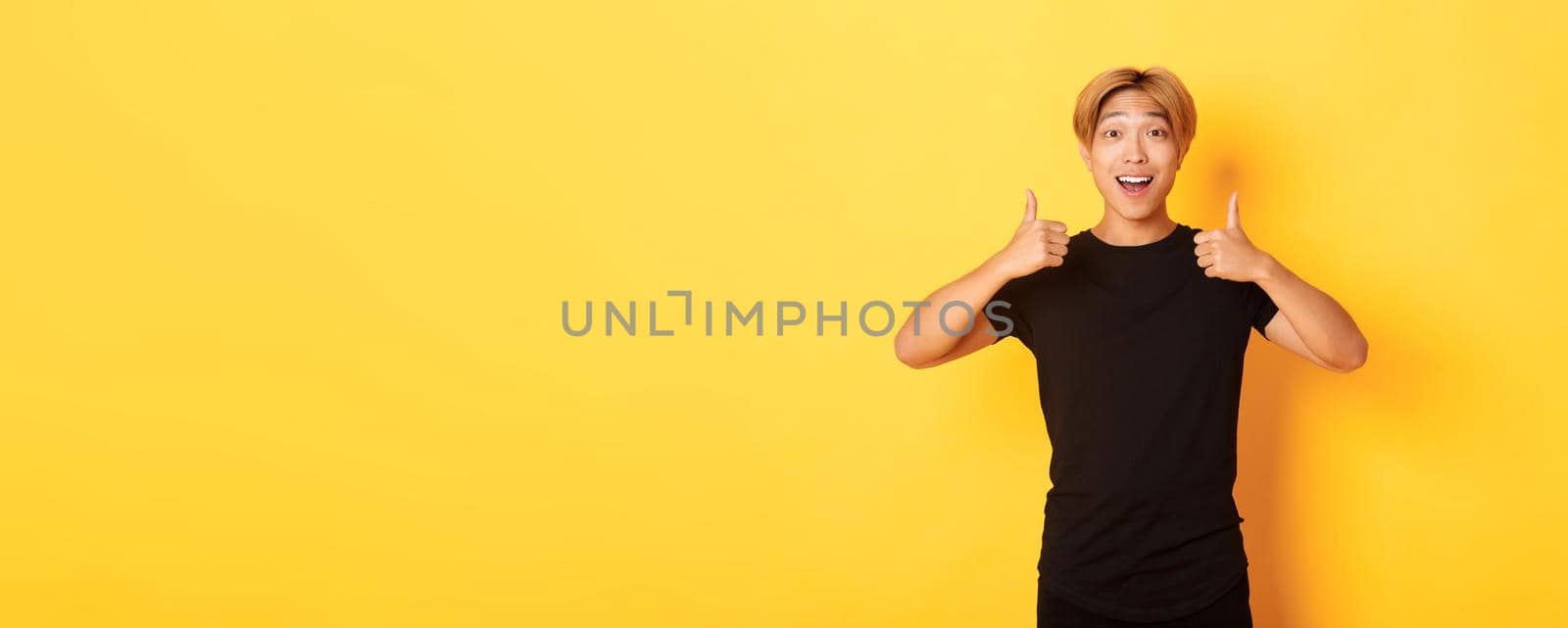Portrait of amazed and pleased asian guy in black outfit, showing thumbs-up in approval, standing excited over yellow background by Benzoix