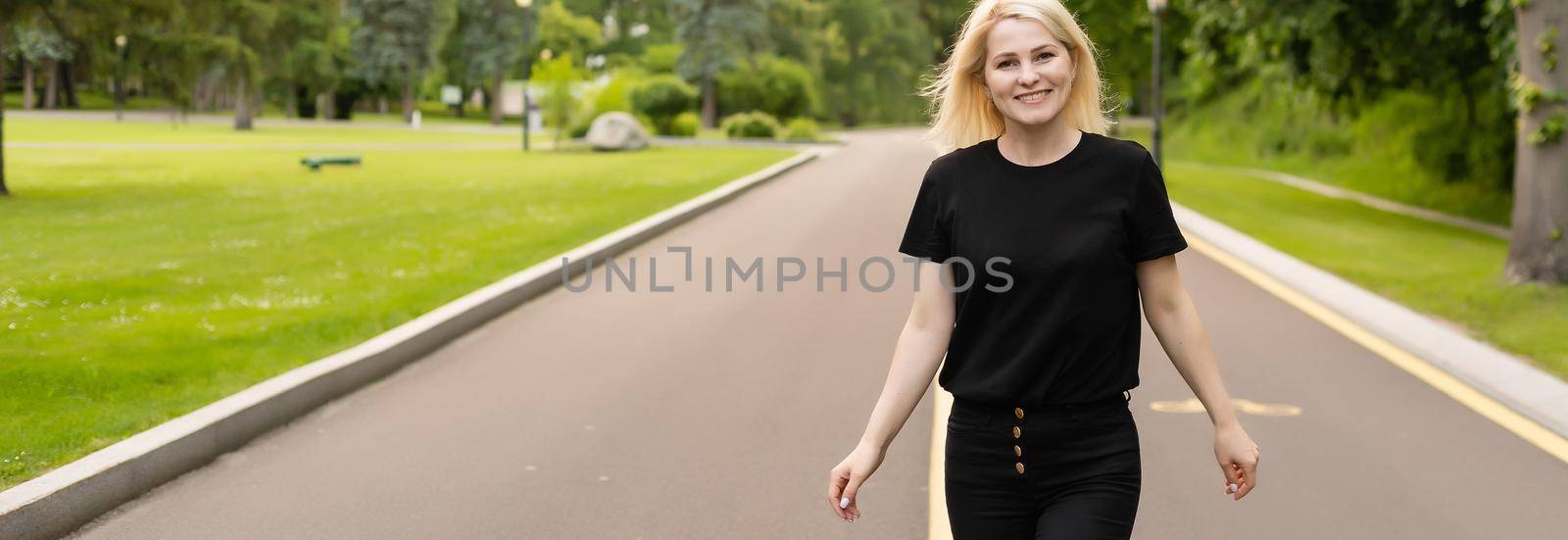 Joyful young lady in casual clothes walking along asphalt road in countryside, hitchhiking for ride outdoors. Lovely millennial woman traveling alone by autostop by Andelov13