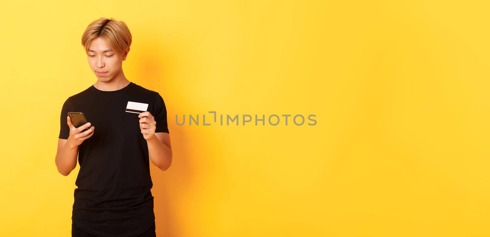 Young handsome asian guy in black t-shirt shopping online, holding credit card and using smartphone, standing yellow background.