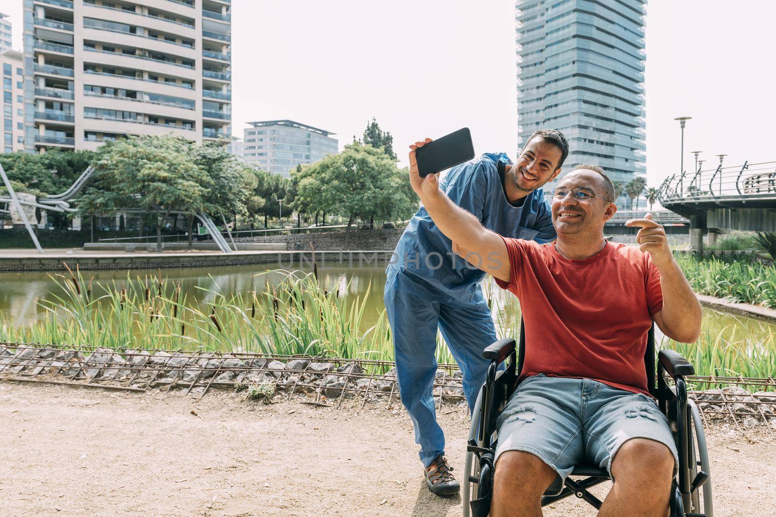 disabled man in a wheelchair taking a selfie with his caretaker during a walk, concept of medical care and rehabilitation of people with disabilities and reduced mobility problems
