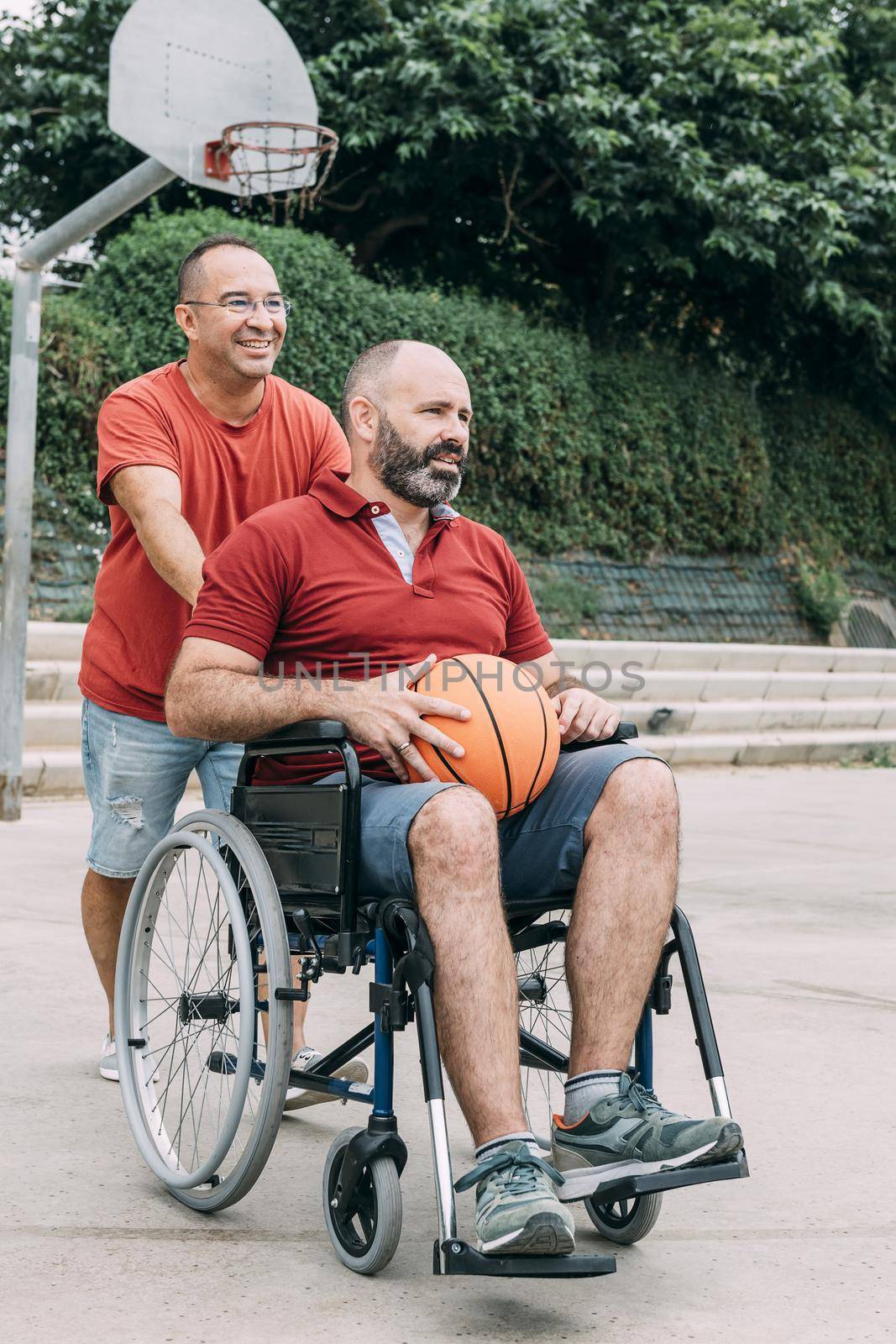 man pushing the wheelchair to play basketball with his handicapped friend, concept of adaptive sports and physical activity, rehabilitation for people with physical disabilities, vertical photo