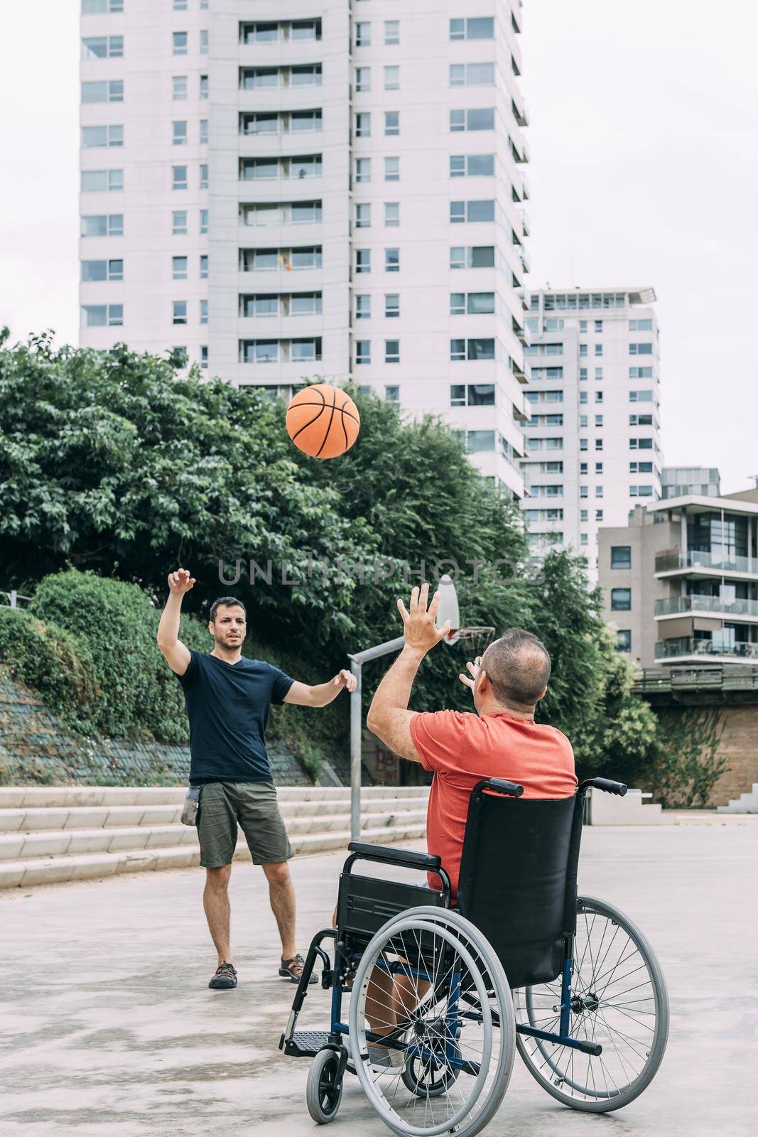 joyful handicapped man in wheelchair playing basketball with a friend with ball, concept of adaptive sports and physical activity, rehabilitation for people with physical disabilities, vertical photo