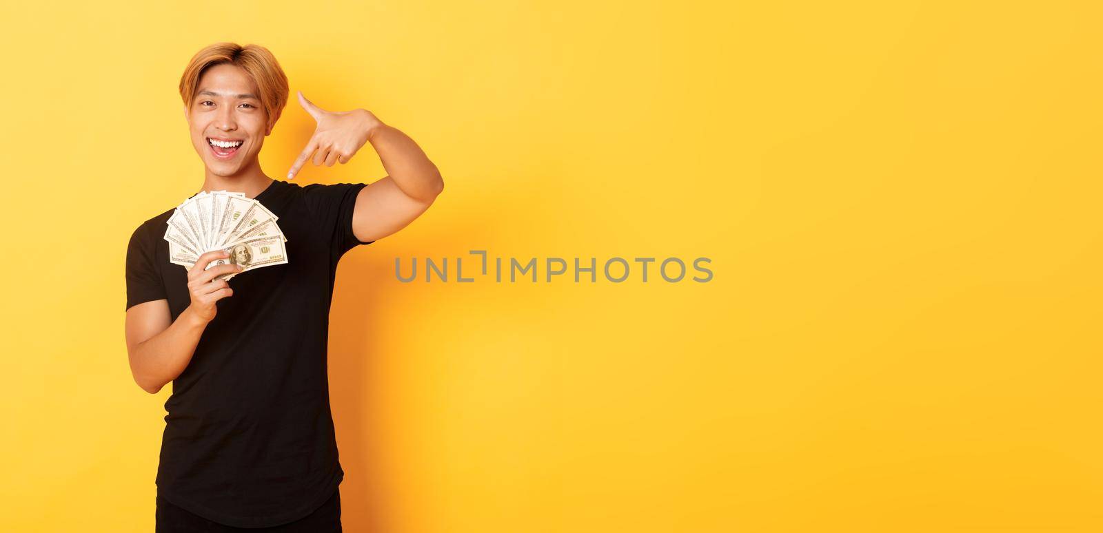 Sassy good-looking korean blond guy, smiling happy and pointing finger at money, winning cash, standing yellow background by Benzoix