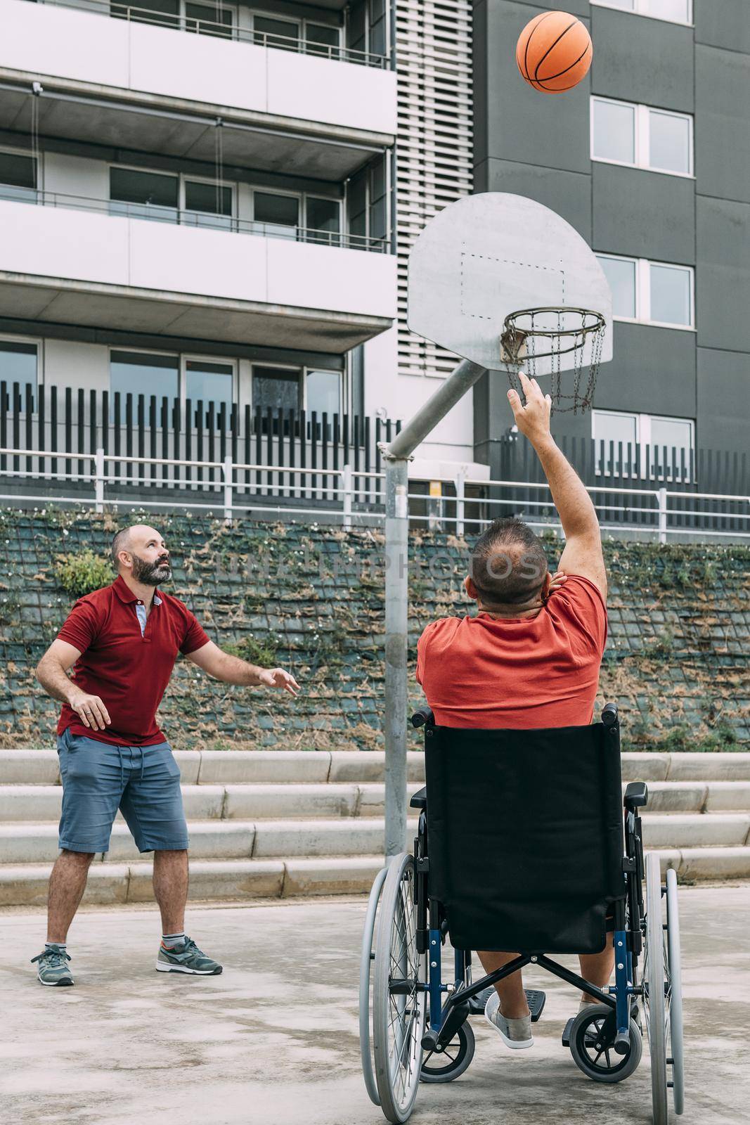 joyful handicapped man in wheelchair throwing the ball to basket with a friend, concept of adaptive sports and physical activity, rehabilitation for people with physical disabilities, vertical photo
