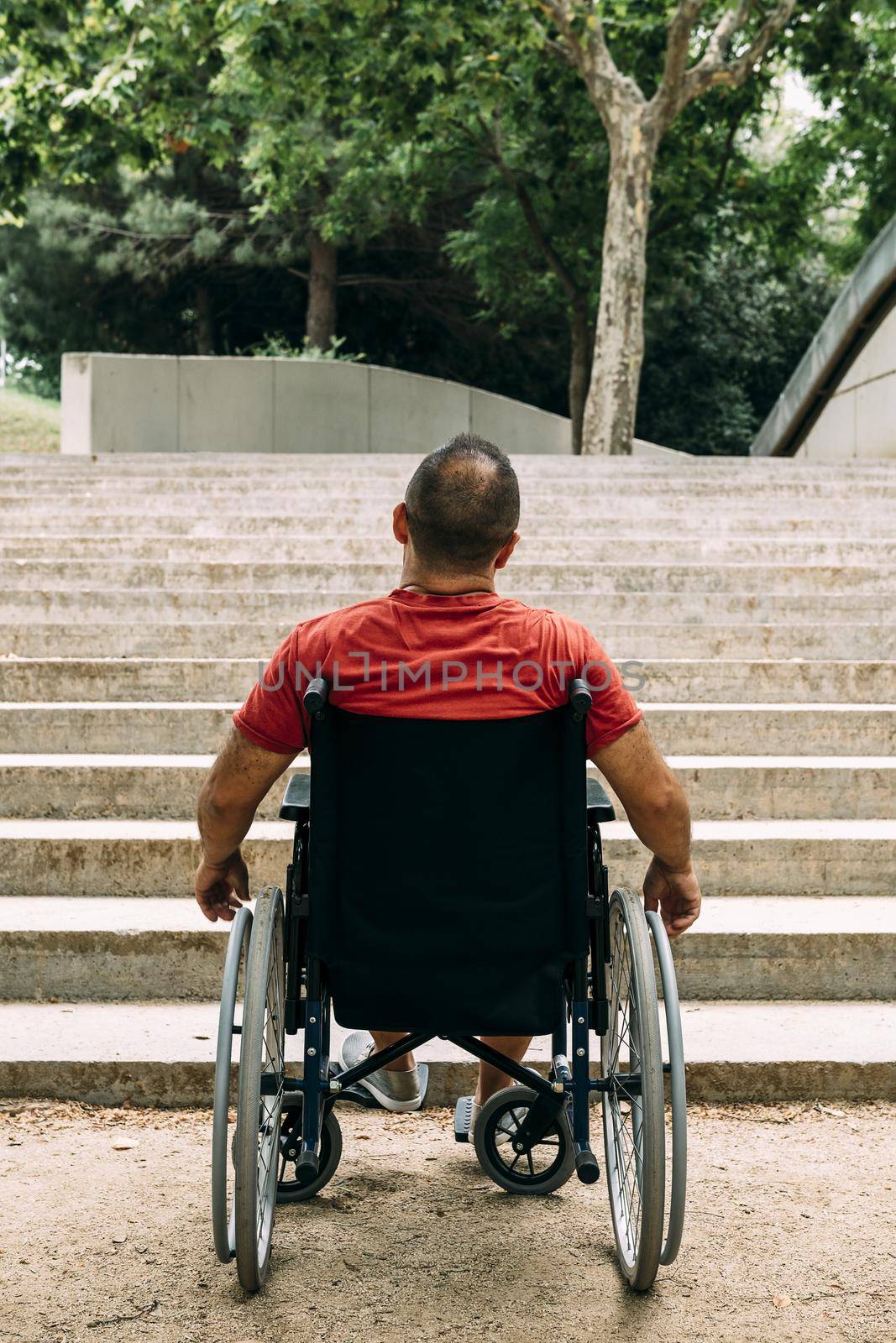vertical photo of a disabled man on wheelchair stopped in front of stairs who can't climb looking up for help, raising awareness of accessibility issues for people with reduced mobility