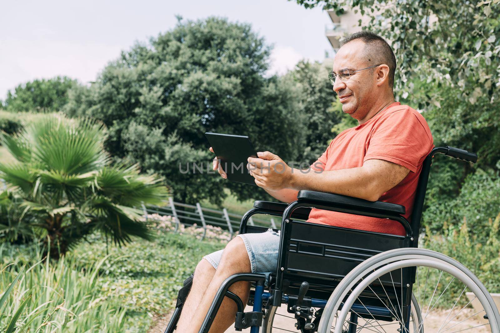 disabled man in wheelchair having fun while working at park using a tablet computer, concept of technological and occupational integration of people with disabilities and reduced mobility problems