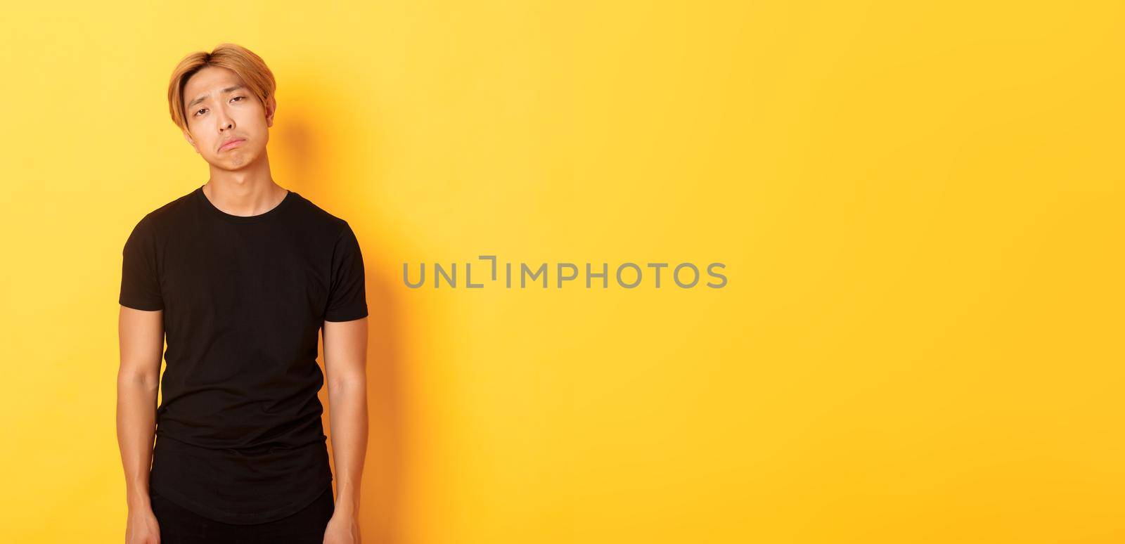 Portrait of exhausted and sad asian guy looking sad, standing drained over yellow background.