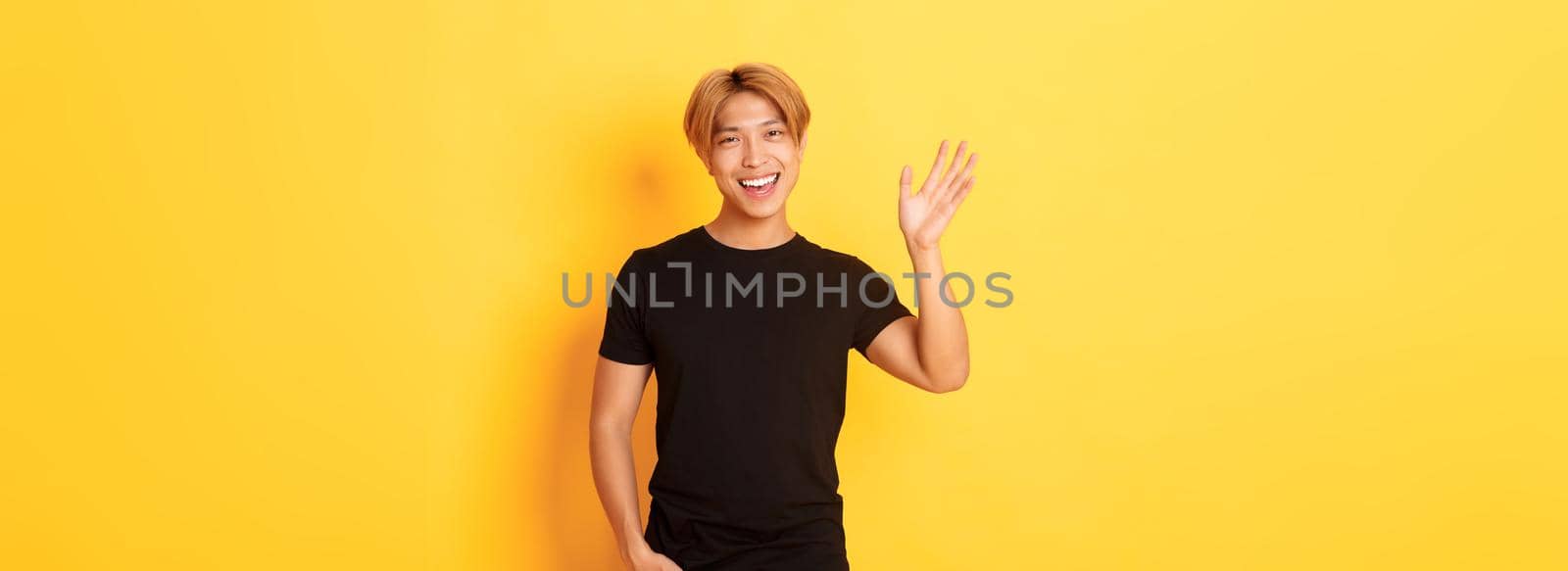 Portrait of handsome friendly asian guy in black outfit, waving hand to say hello and smiling, greeting someone, standing yellow background.