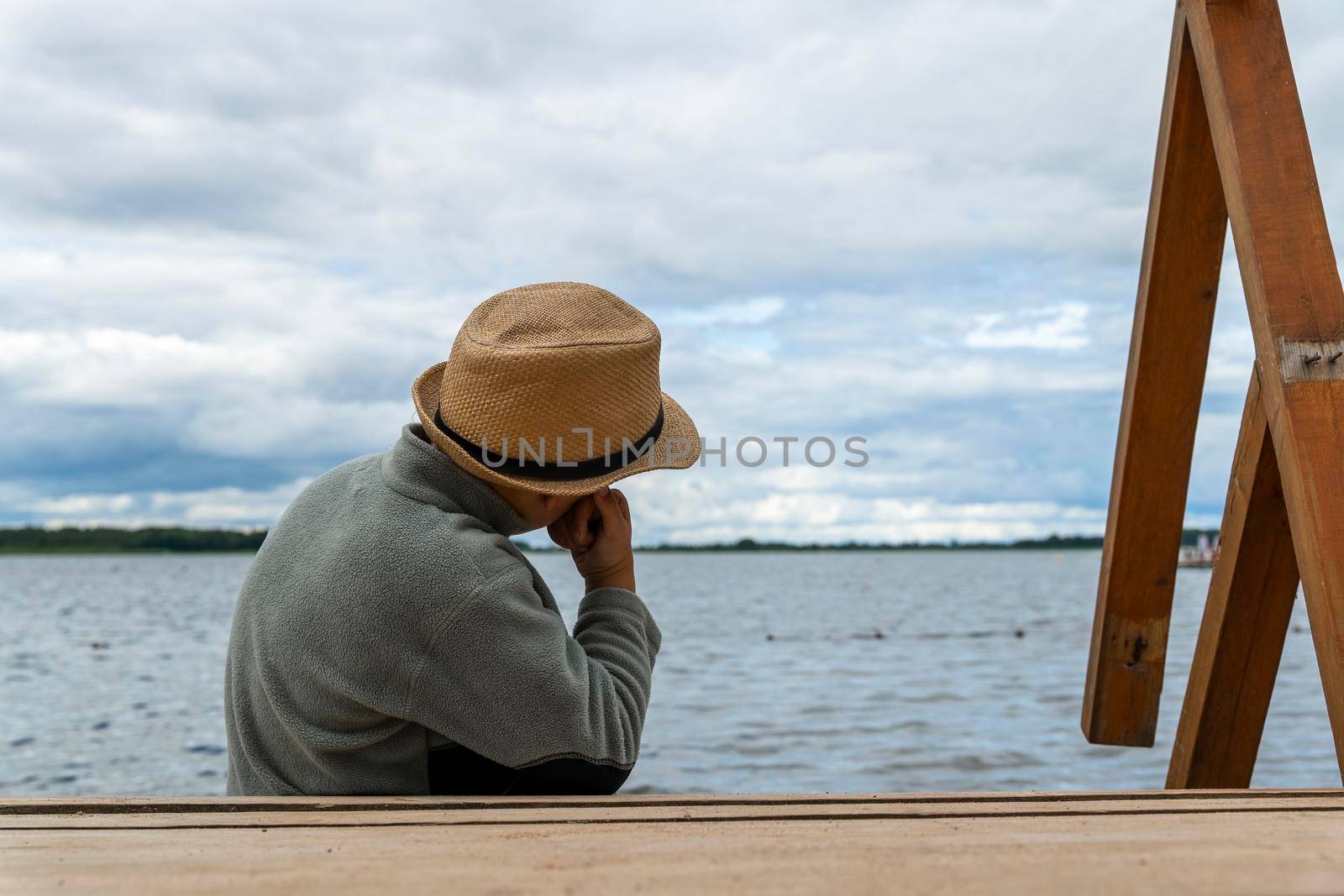 a boy in a hat sits on the shore of a lake and looks into the distance by audiznam2609