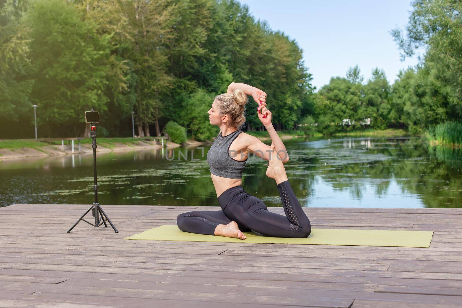 A slender beautiful woman in a gray top and leggings, on a wooden platform by a pond in a park in summer, does yoga, doing stretching, on a green sports mat, in front of her smartphone on a tripod.