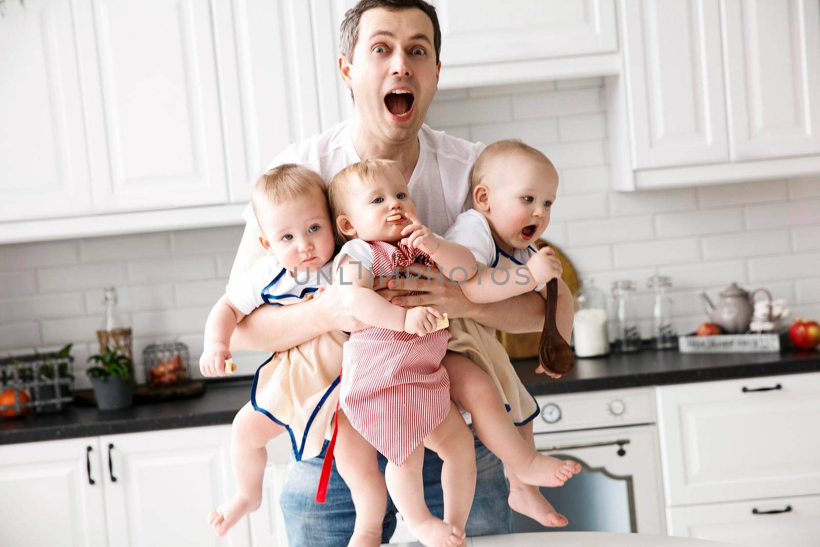 A man holds and hugs three babies in his arms. Happy father of triplets in front of a white kitchen. The concept of a happy family, eco, healthy food. Father on parental leave