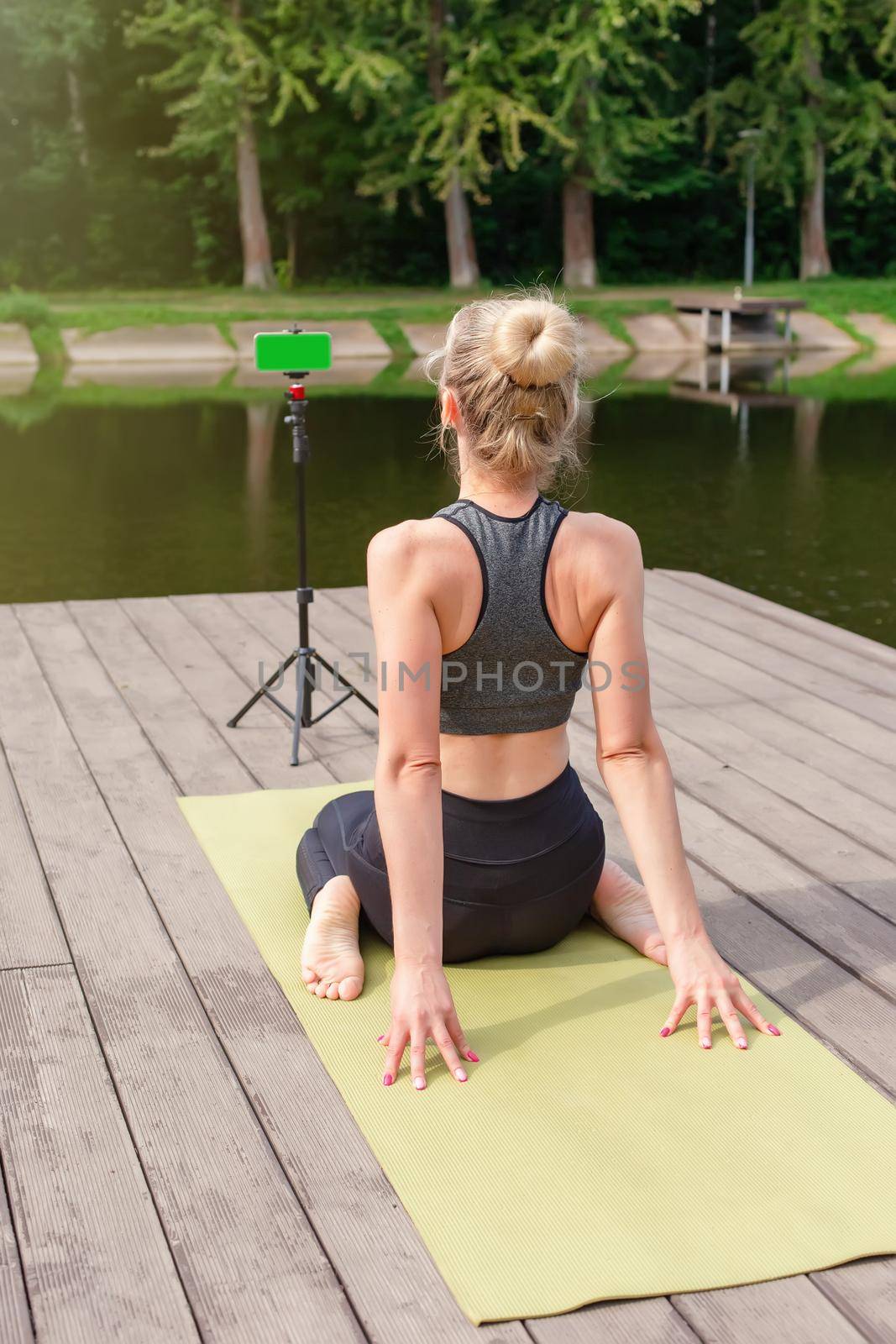 A woman in a gray sportswear, sits on a wooden platform by a pond in the park in summer, does yoga, performs exercises, on a sports mat, in front of her smartphone on a tripod. Vertical
