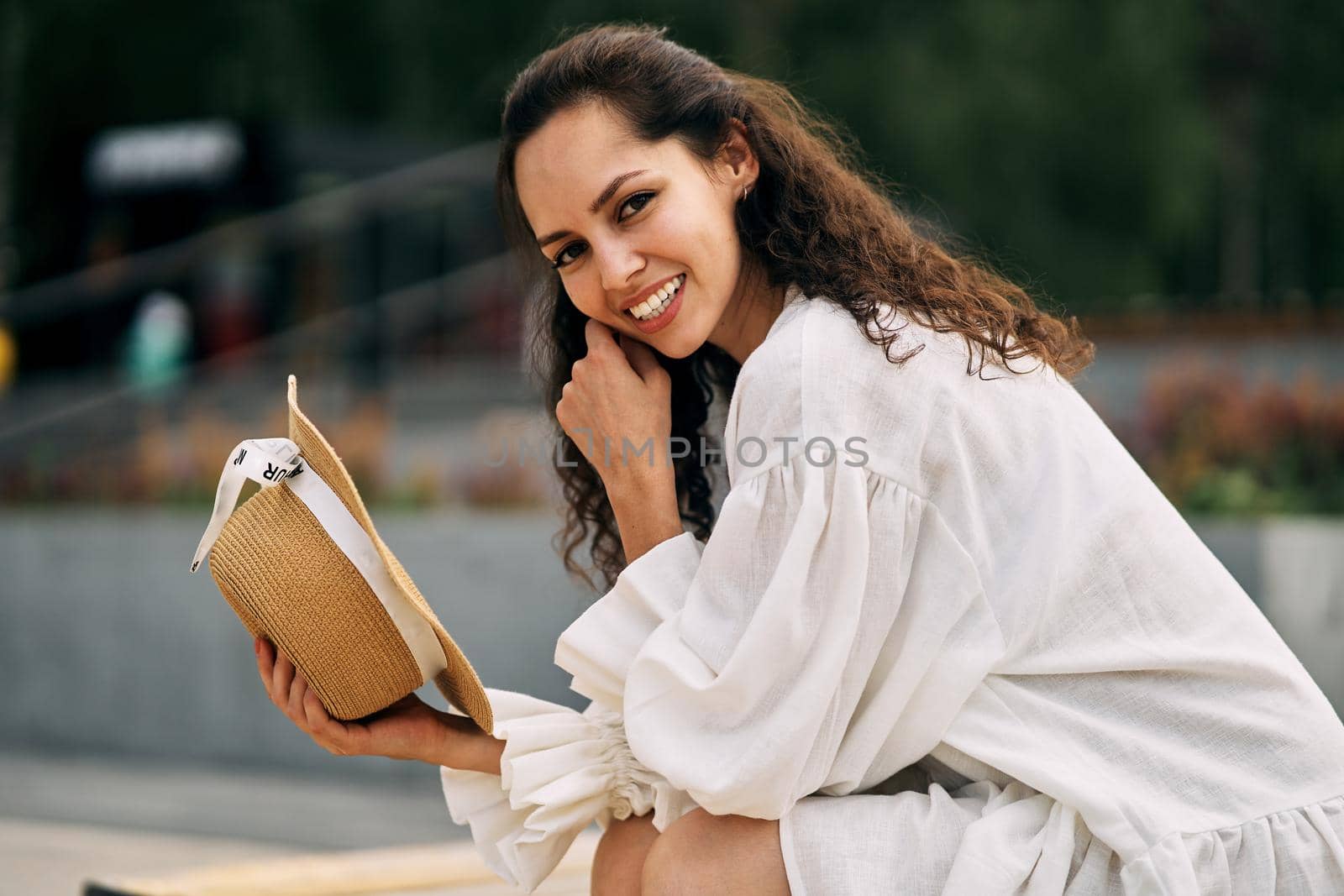 Smiling curly brunette girl in a white dress sitting with a hat in her hand by driver-s
