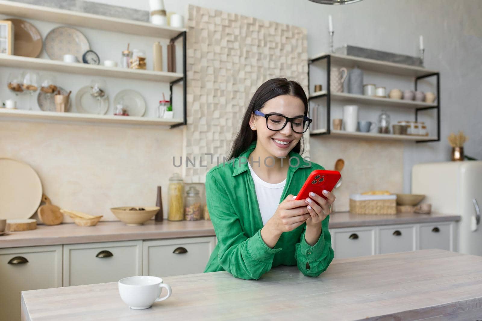 In love and happy young beautiful woman sends and receives love messages on the red phone from a loved one, from a boyfriend. Standing in the kitchen at home in glasses and a green shirt.