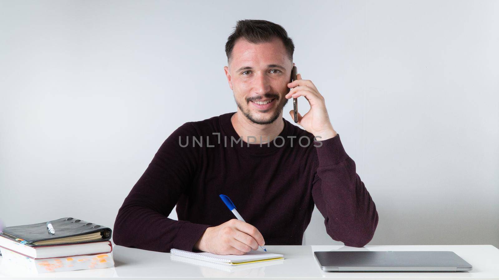 A man talks on the phone and takes notes in a notebook at an office desk. by voktybre