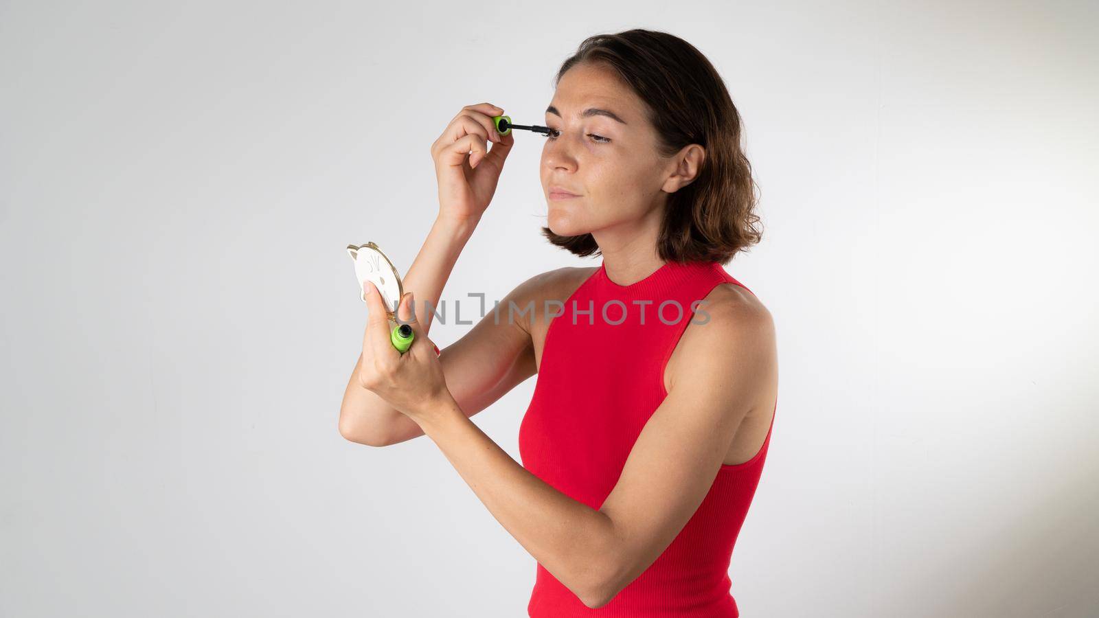 A woman dyes her eyelashes with mascara and looks in the mirror on a white background. High quality photo