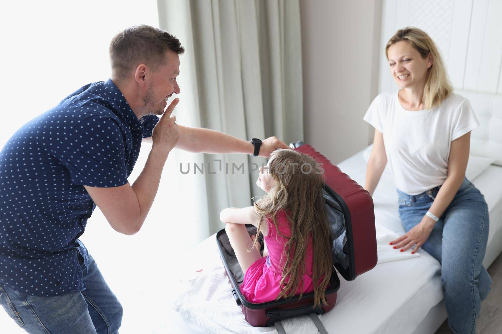 Cheerful dad and mom play hide and seek with child and suitcase. Dad asks child to sit quietly in suitcase