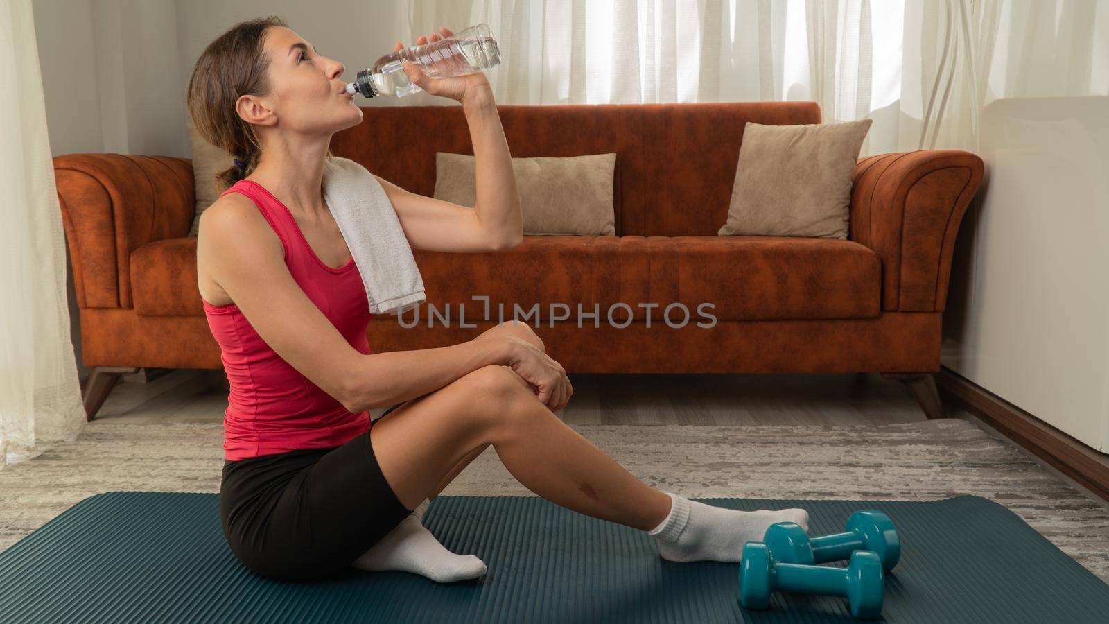 A woman after a home exercise drinks water on a mat next to dumbbells by voktybre