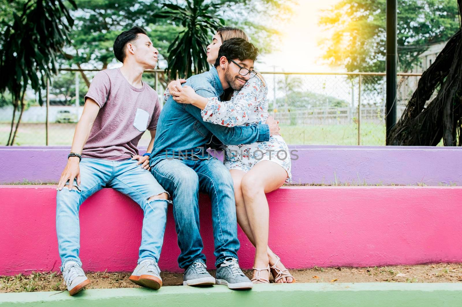 Unfaithful girl sitting hugging her boyfriend and secretly kissing another man. Unfaithful girlfriend hugging her boyfriend in a park secretly kissing another man, Couple infidelity concept
