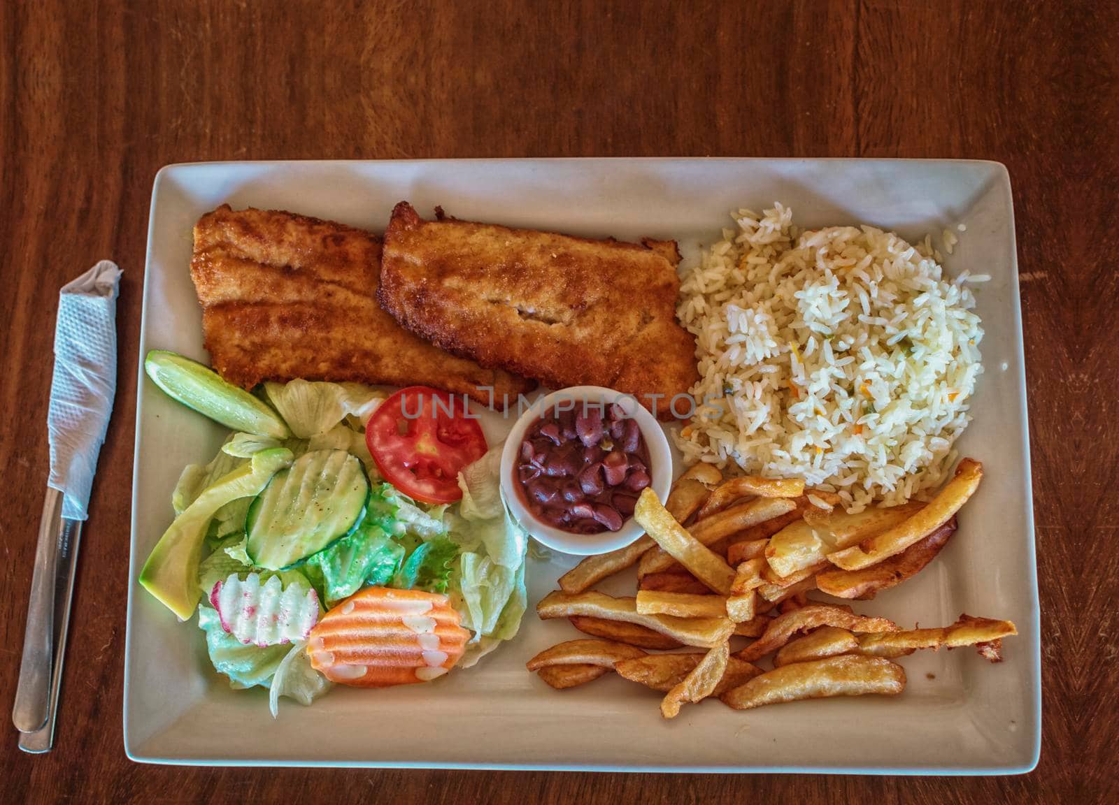 Plate of fried fish fillet with salad and french fries on wooden table, Top view of delicious fried fish fillet with french fries, rice and salad served on wooden table. Gourmet food plate