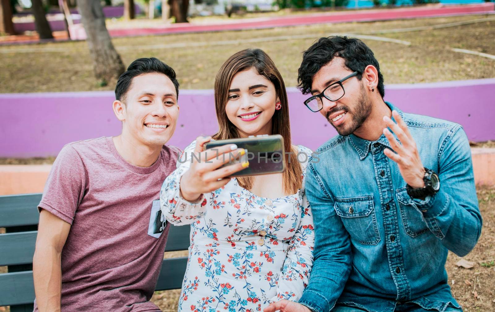 Close up of three friends taking a selfie sitting in the park, Three smiling friends sitting in the park taking a selfie. Front view of three happy friends taking a selfie while sitting on a bench