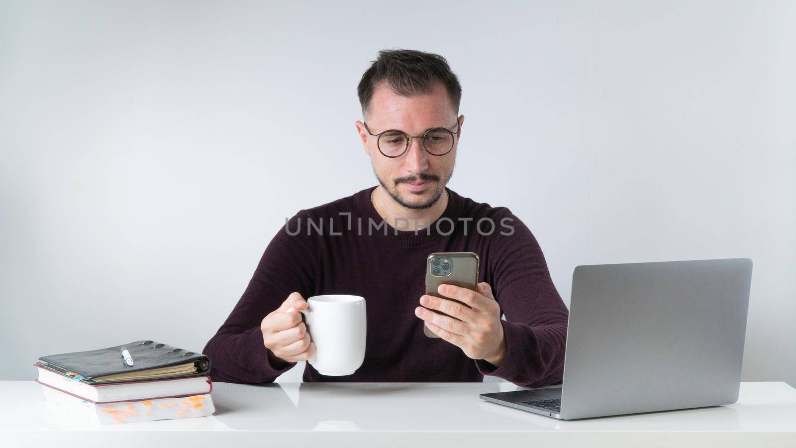 A man with glasses with a phone in his hand and a cup of coffee on his desk with a laptop by voktybre