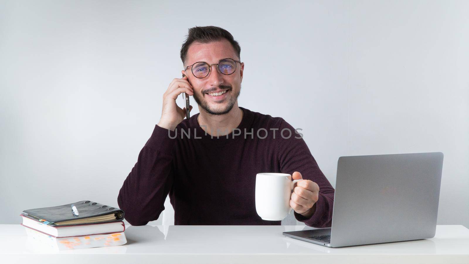 A man works at a laptop with a phone in his hand and a cup of coffee by voktybre