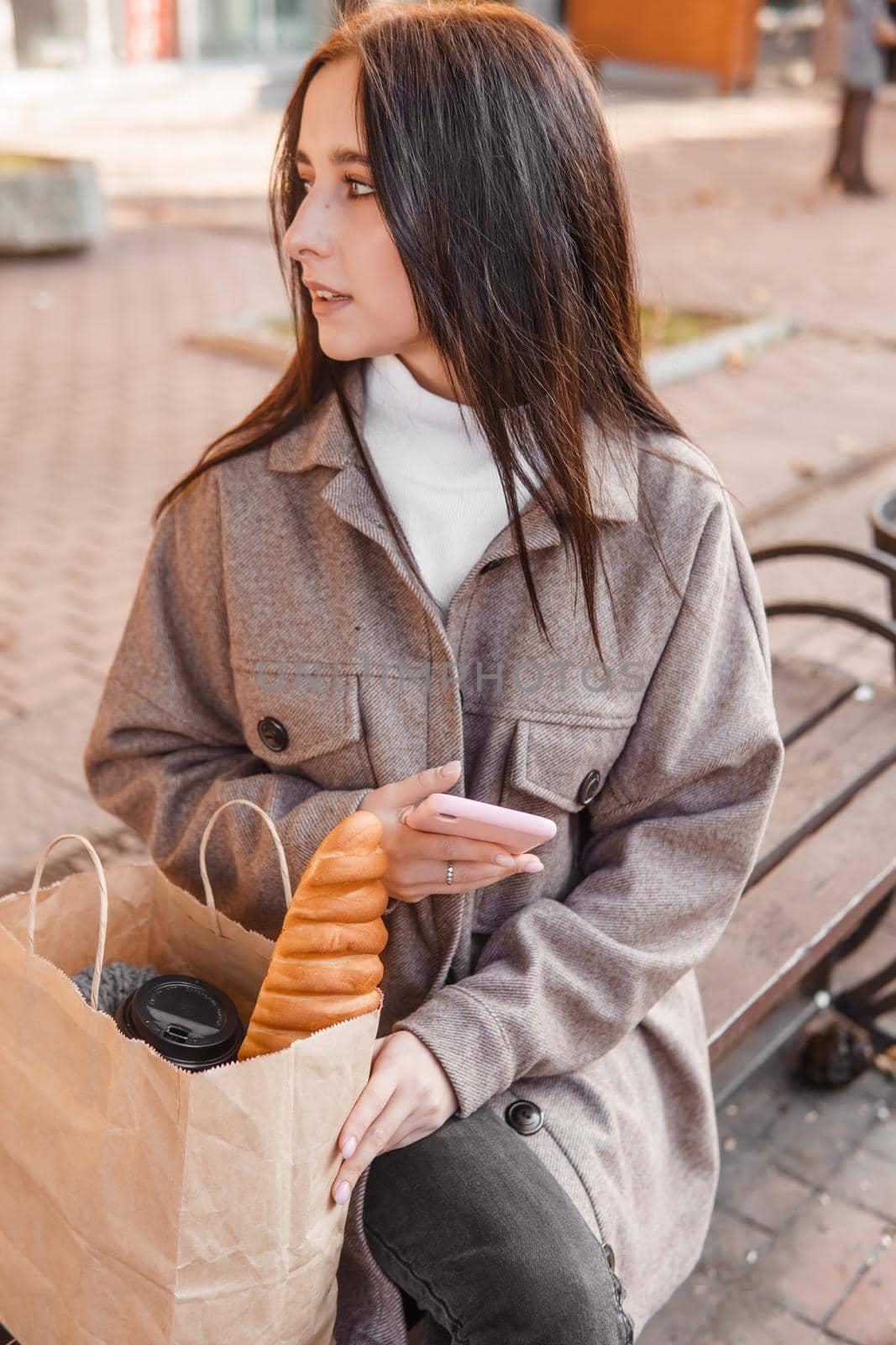 A stylish brunette woman walks around the autumn city. The brunette is sitting on a bench outside with a glass of coffee and a phone, holding a craft bag with a baguette.