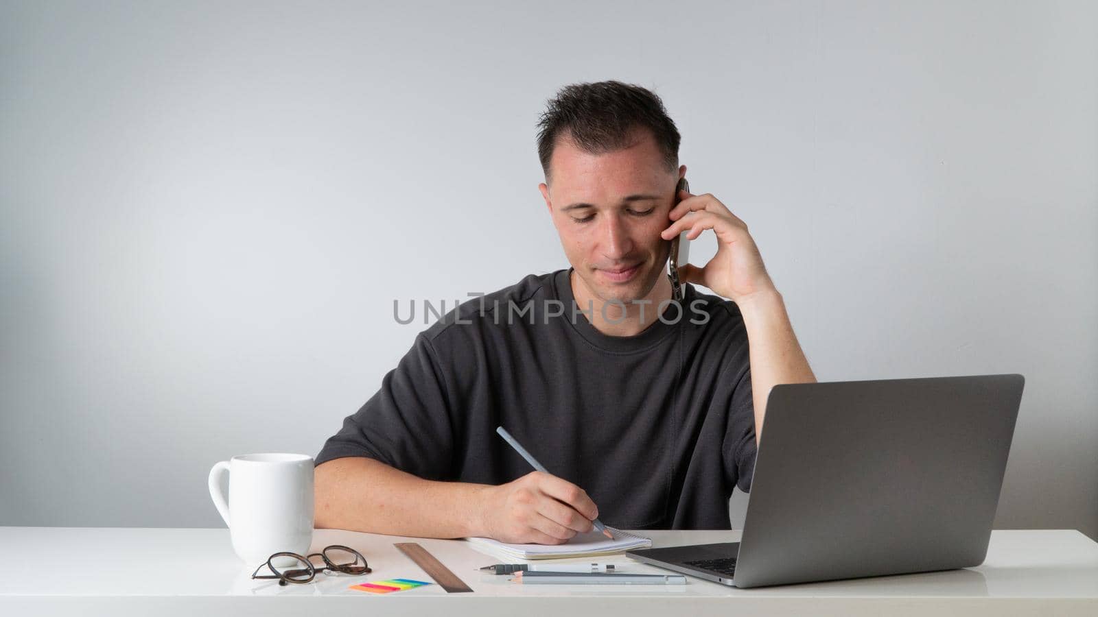 Male student writes a lecture in a notebook and talks on the phone with a laptop by voktybre