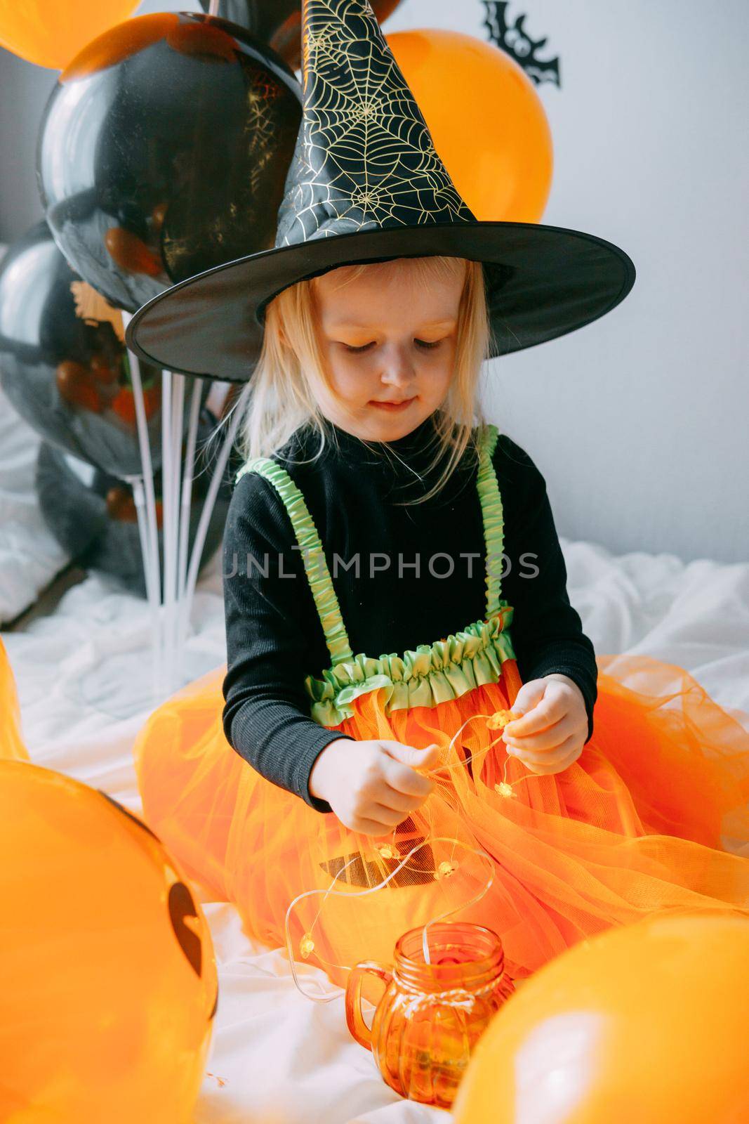 Children's Halloween - a girl in a witch hat and a carnival costume with airy orange and black balloons at home. Ready to celebrate Halloween.