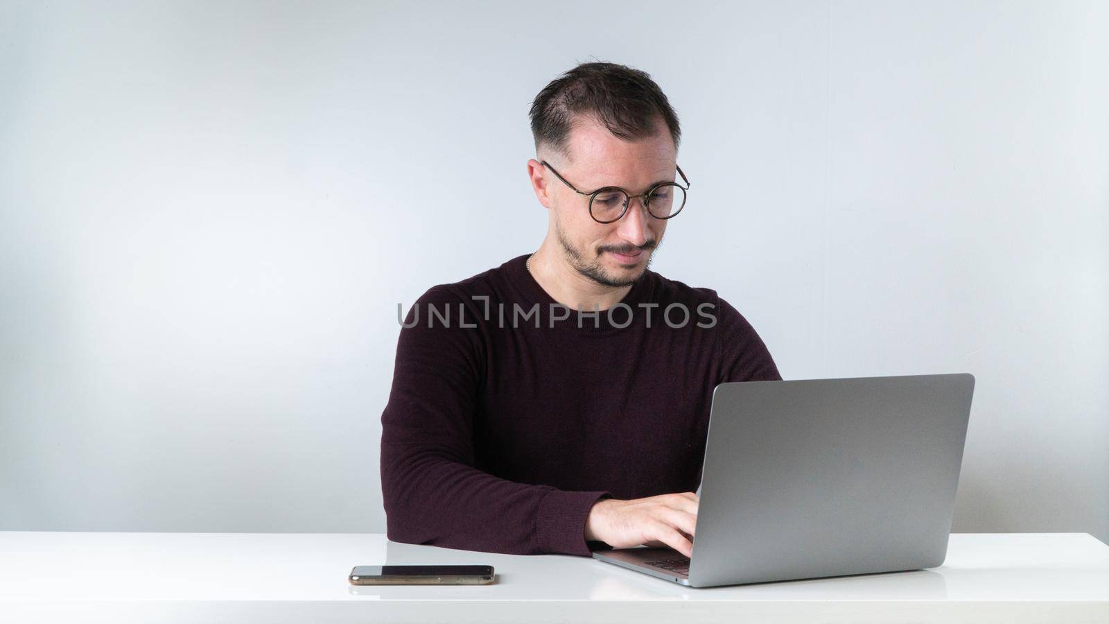 A man with glasses with a beard and mustache types on a laptop at his desk on a white background. High quality photo