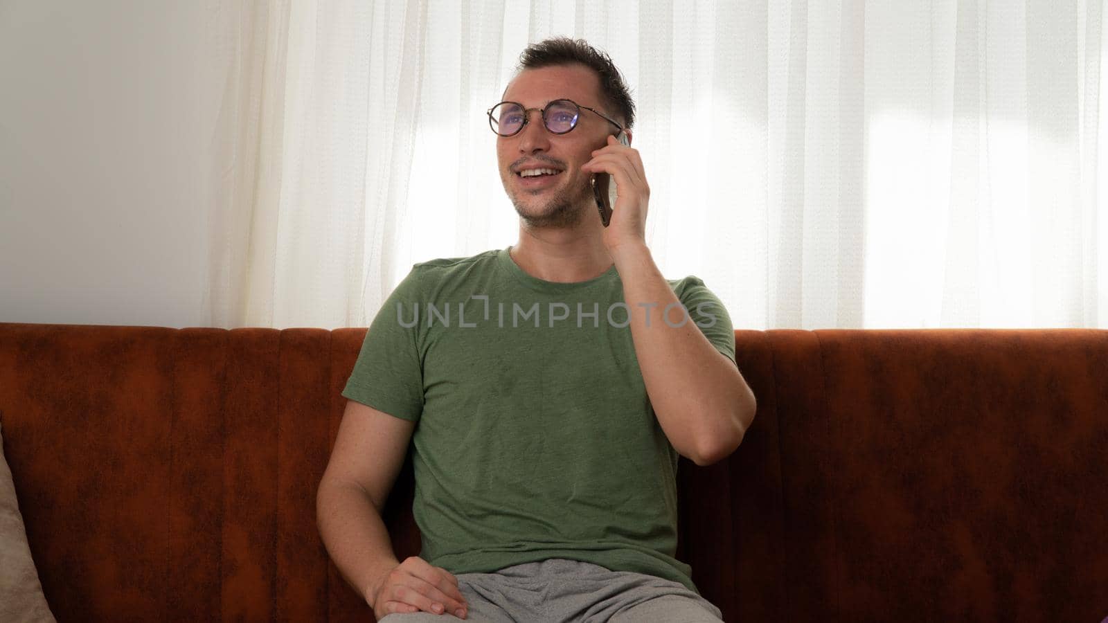 A man happily talks on the phone, communicates at home sitting on the couch by voktybre