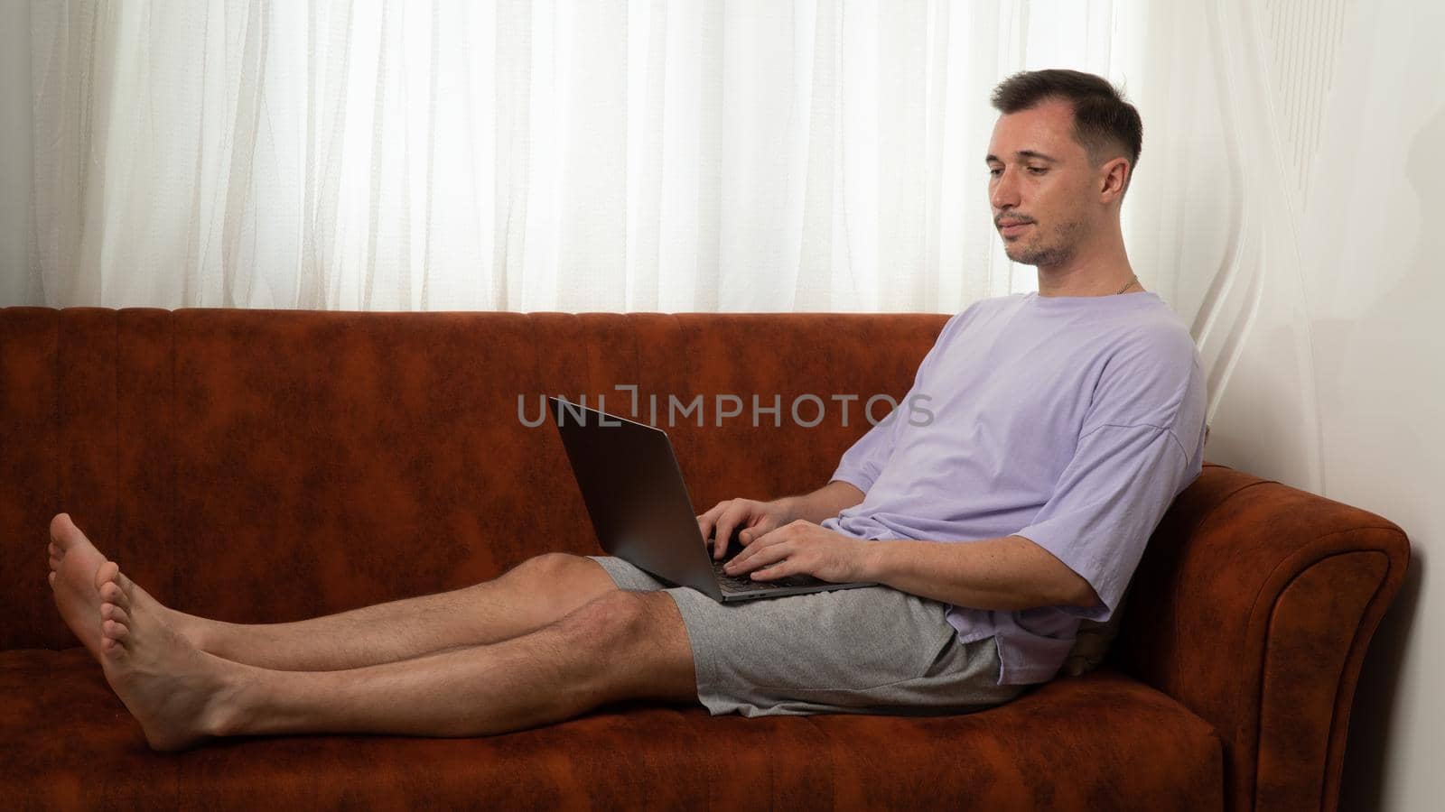 A man lies on the couch and types in a laptop, working at the computer at home by voktybre