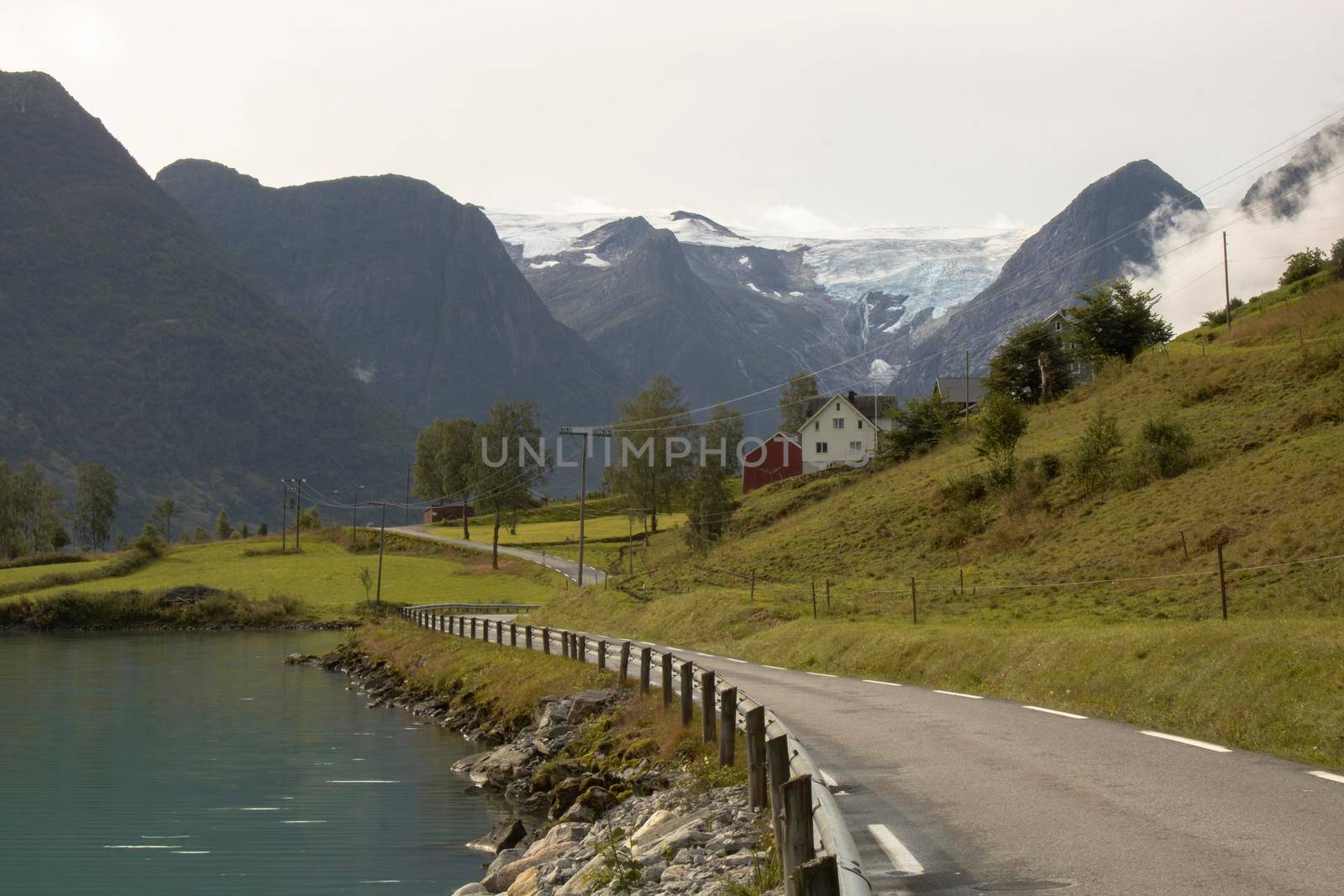 Beautiful landscape showing a road next to a fjord and a house and some snowy mountains in the background in Norway