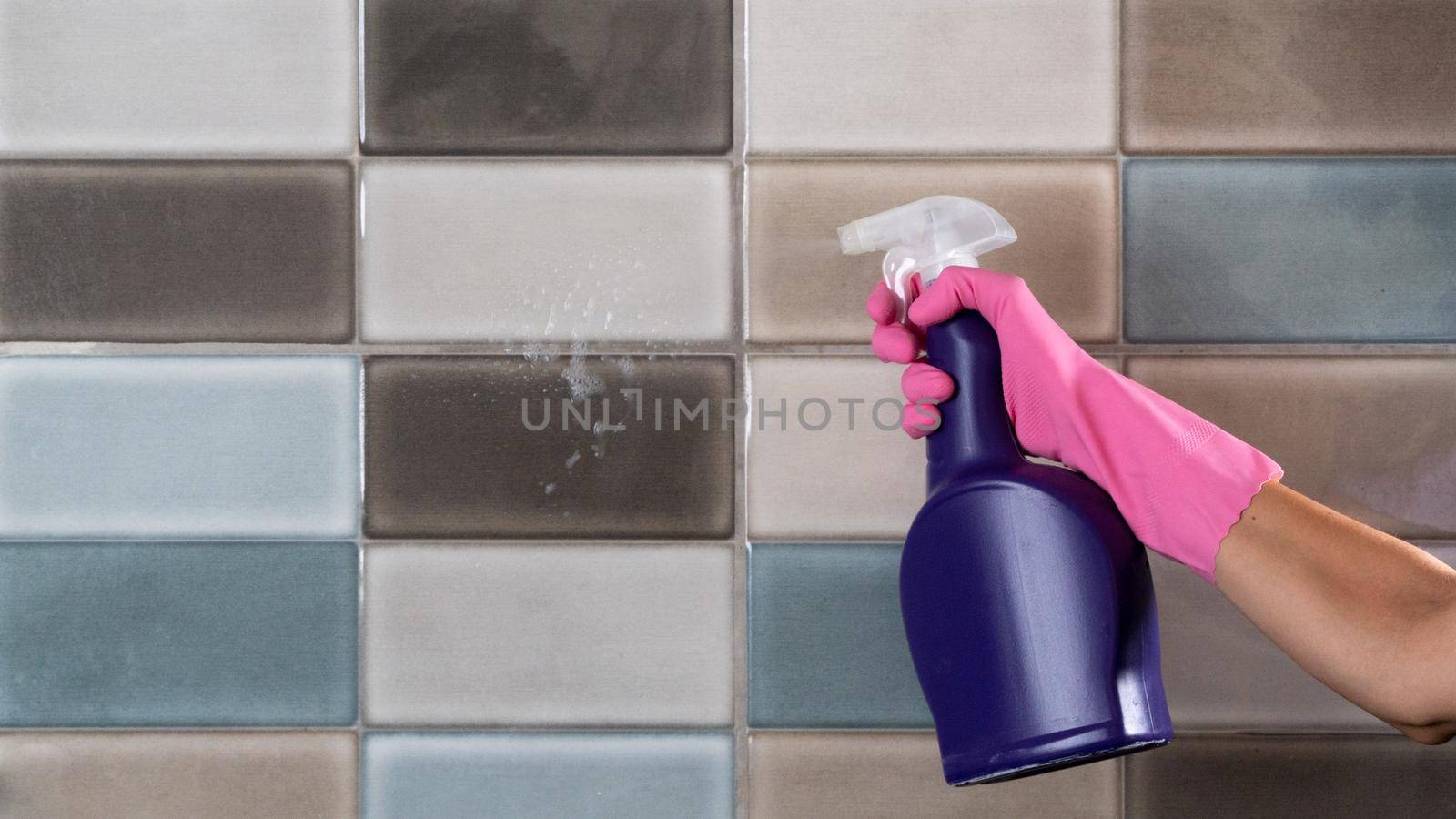 A woman in rubber gloves washes tiles in the kitchen or bathroom with a sponge and sprays detergent