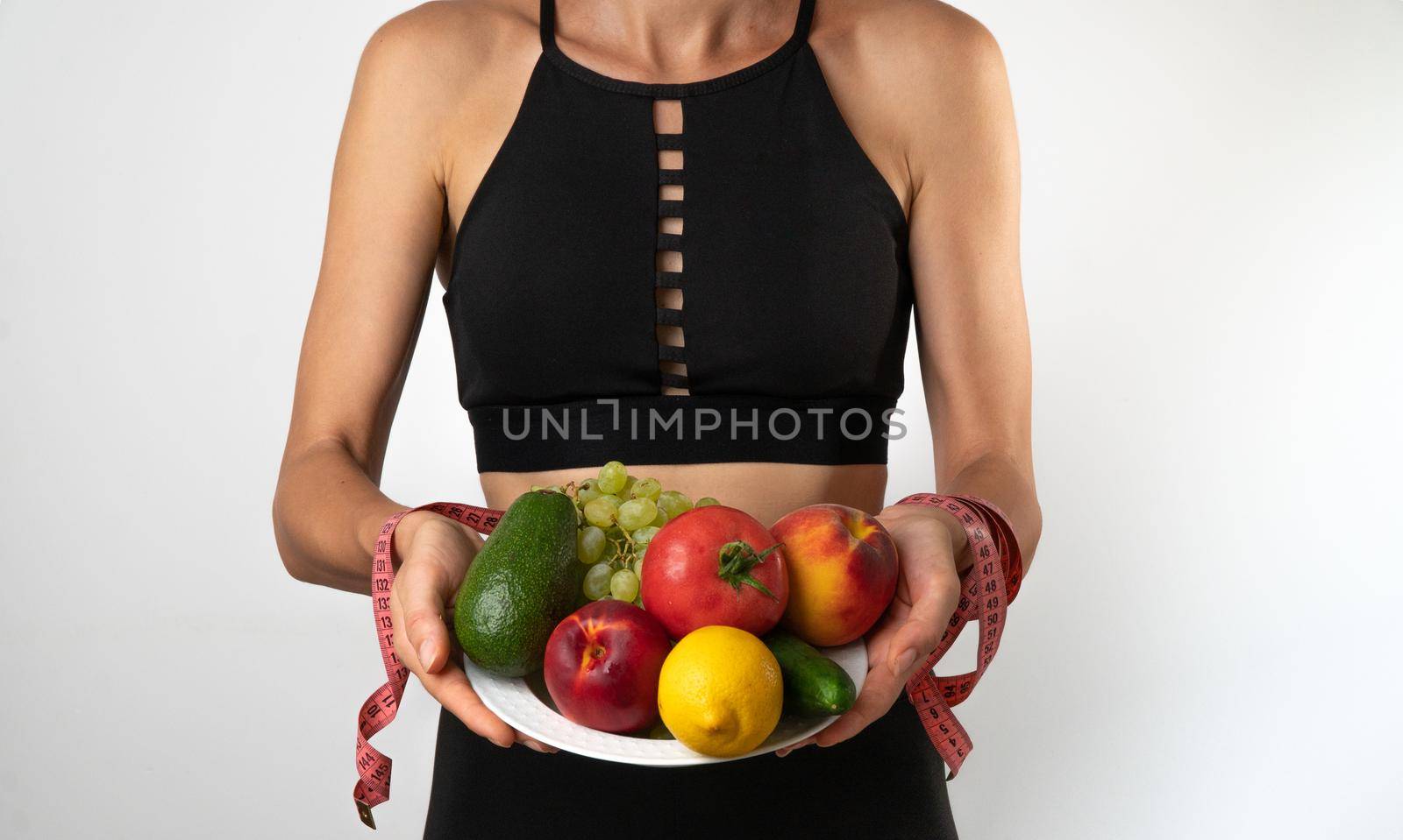 A woman in a sports uniform holds a plate with fruits and vegetables and measuring tape in her hands, trainer nutritionist. High quality photo