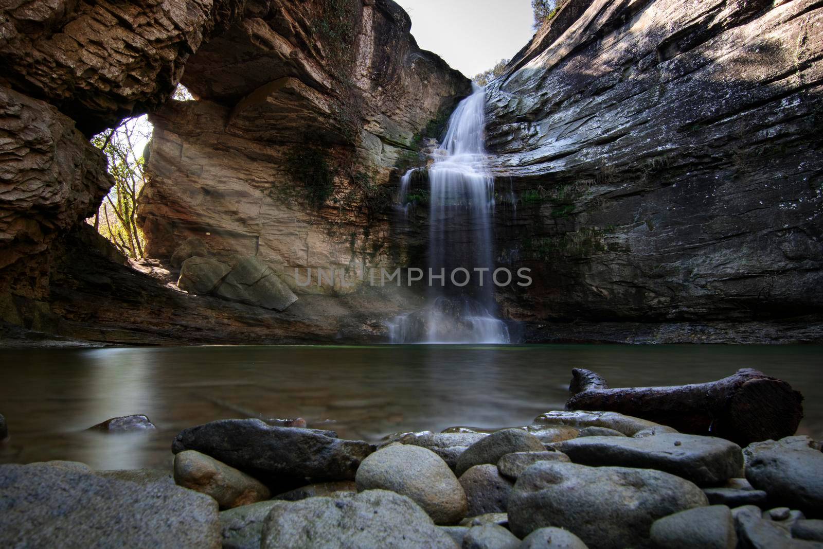 Rays of sunlight entering through a hole in a rock and a waterfall falling on a lake with stones and a big branch in the foreground in a place called Foradada