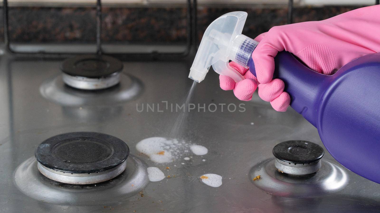 A hand in a pink glove sprays detergent on a dirty gas stove for cleaning by voktybre