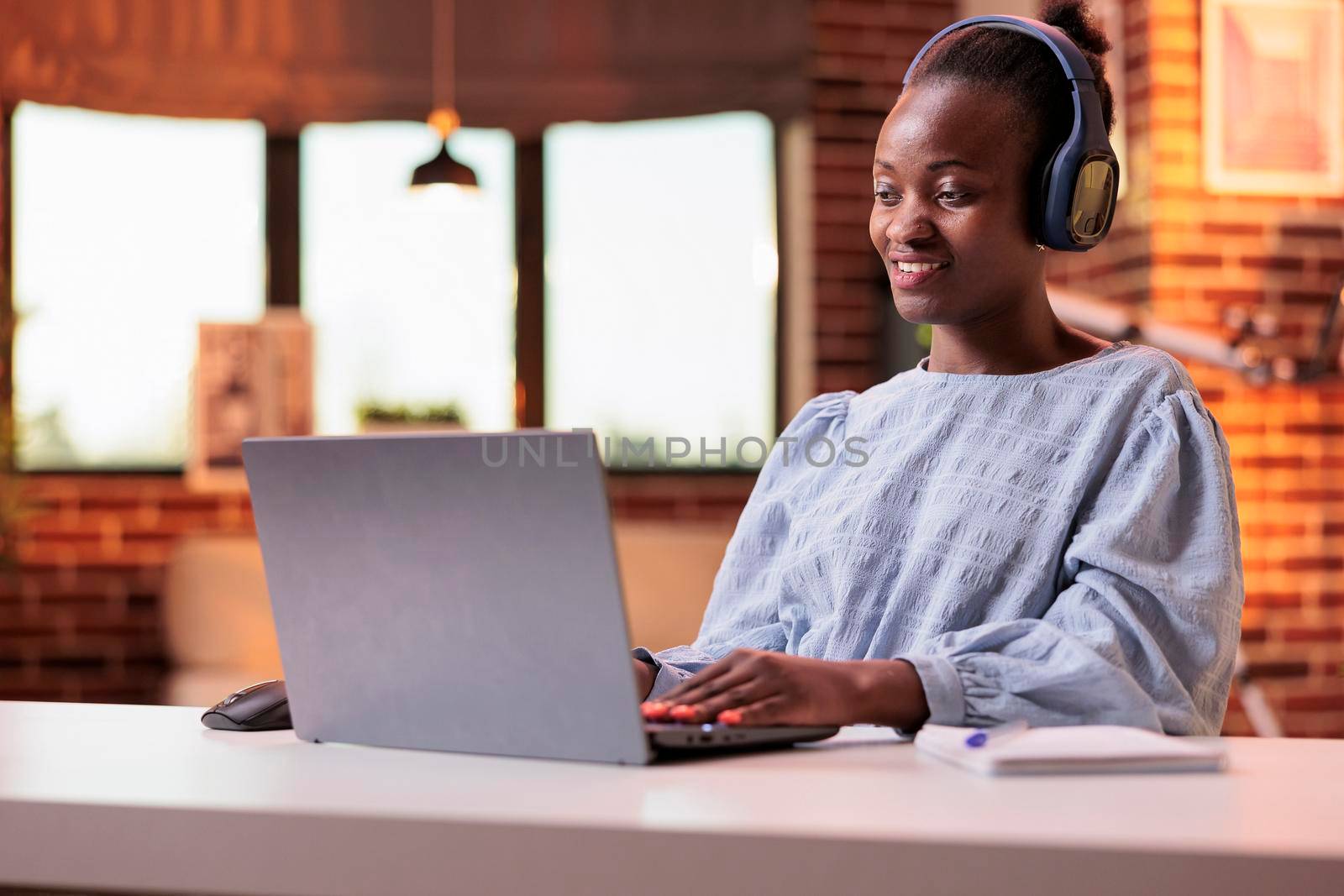 Smiling student in headphones attending online classes on laptop at home. African american woman watching educational video in modern room with big windows and beautiful warm sunset light