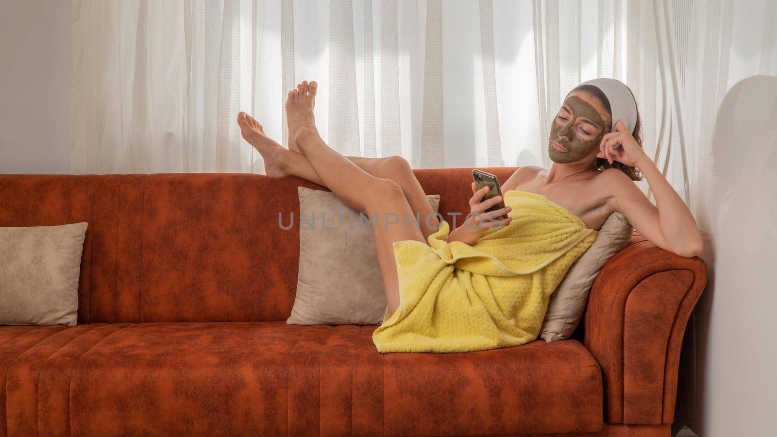 A woman on the couch in a towel waits for the clay mask on her face to dry, looks at the phone by voktybre