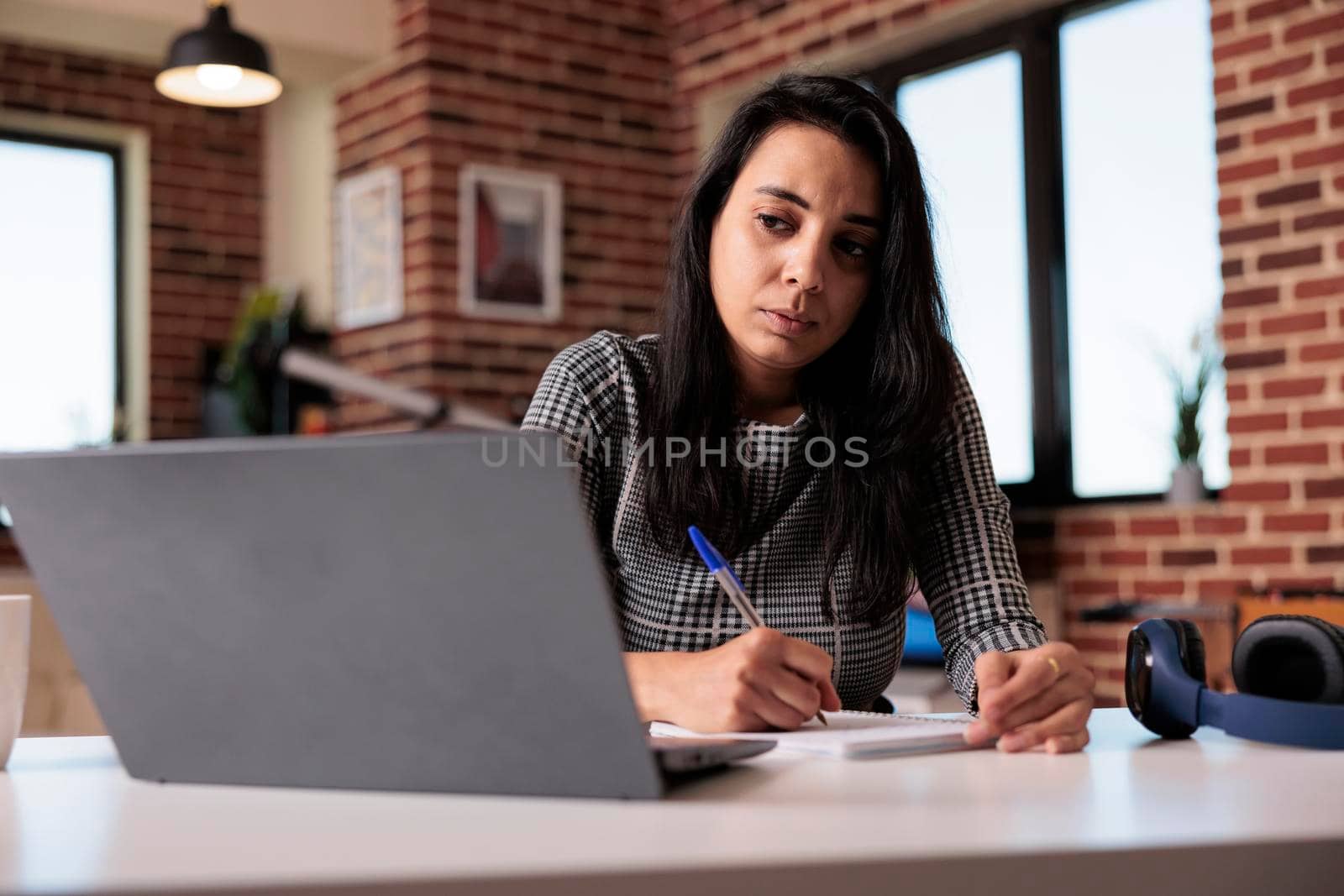 Indian business woman taking paperwork notes on notebook, writing information report or school class lesson. Working on laptop with textbook to do research online and plan web presentation.