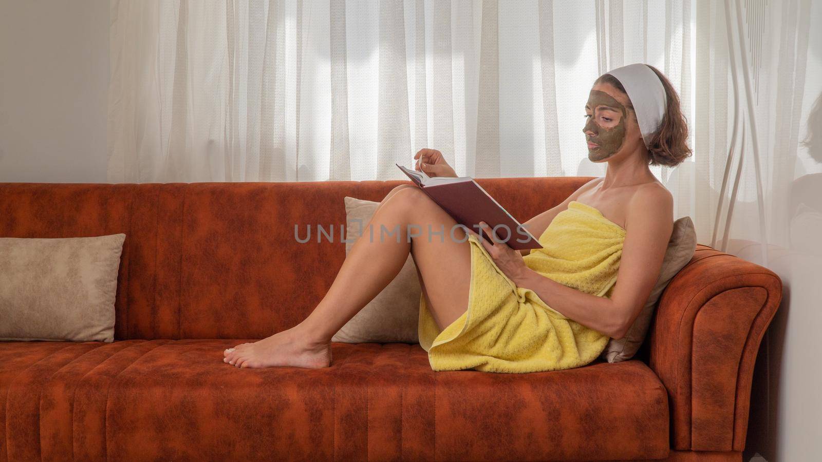 A woman with a mask on her face and in a towel reads a book on the couch - face care at home by voktybre