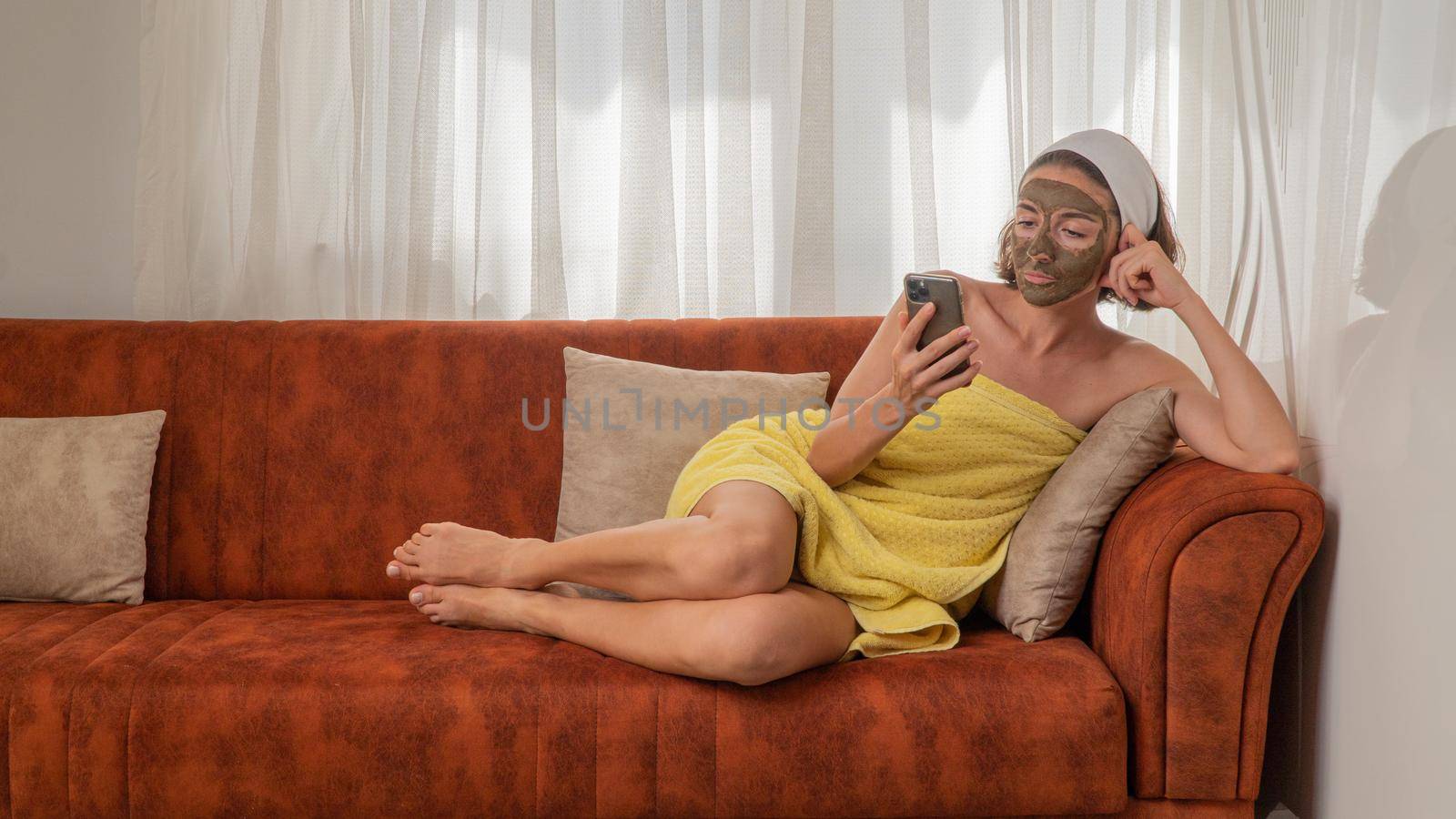 A woman with a clay mask on her face in a towel lies on a sofa with a smartphone. High quality photo