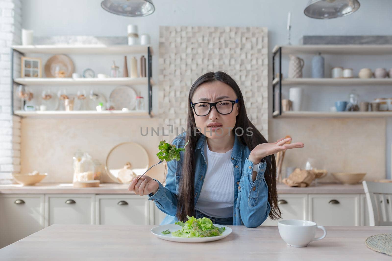 Portrait of young dissatisfied Asian woman eating salad at home and looking at camera.