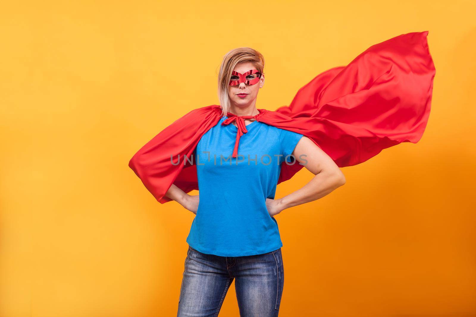 Young woman in superheros costume standing proudly with her red cape in the air over yellow background. Super powers.