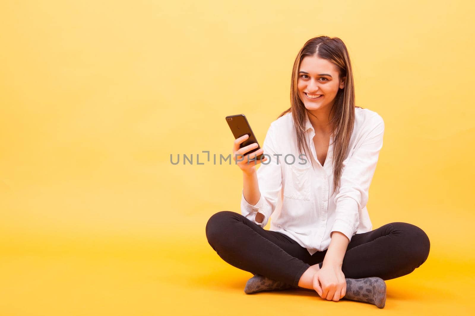 Beautiful girl smiling her phone while sitting down over yellow background. Facial expression