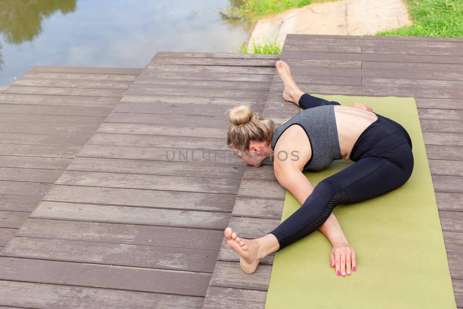 A slender woman in a gray sportswear does yoga in summer on a wooden platform by a pond in the park, stretching . Copy space.