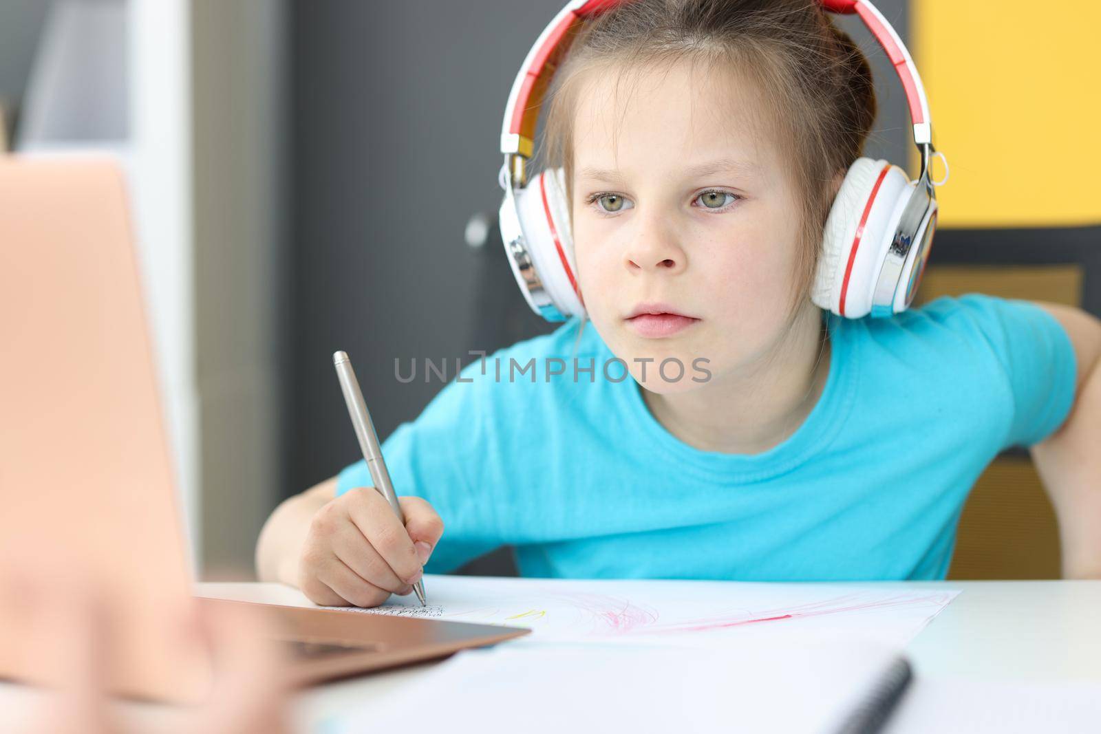 Little girl schoolgirl in headphones sitting in front of laptop at home by kuprevich