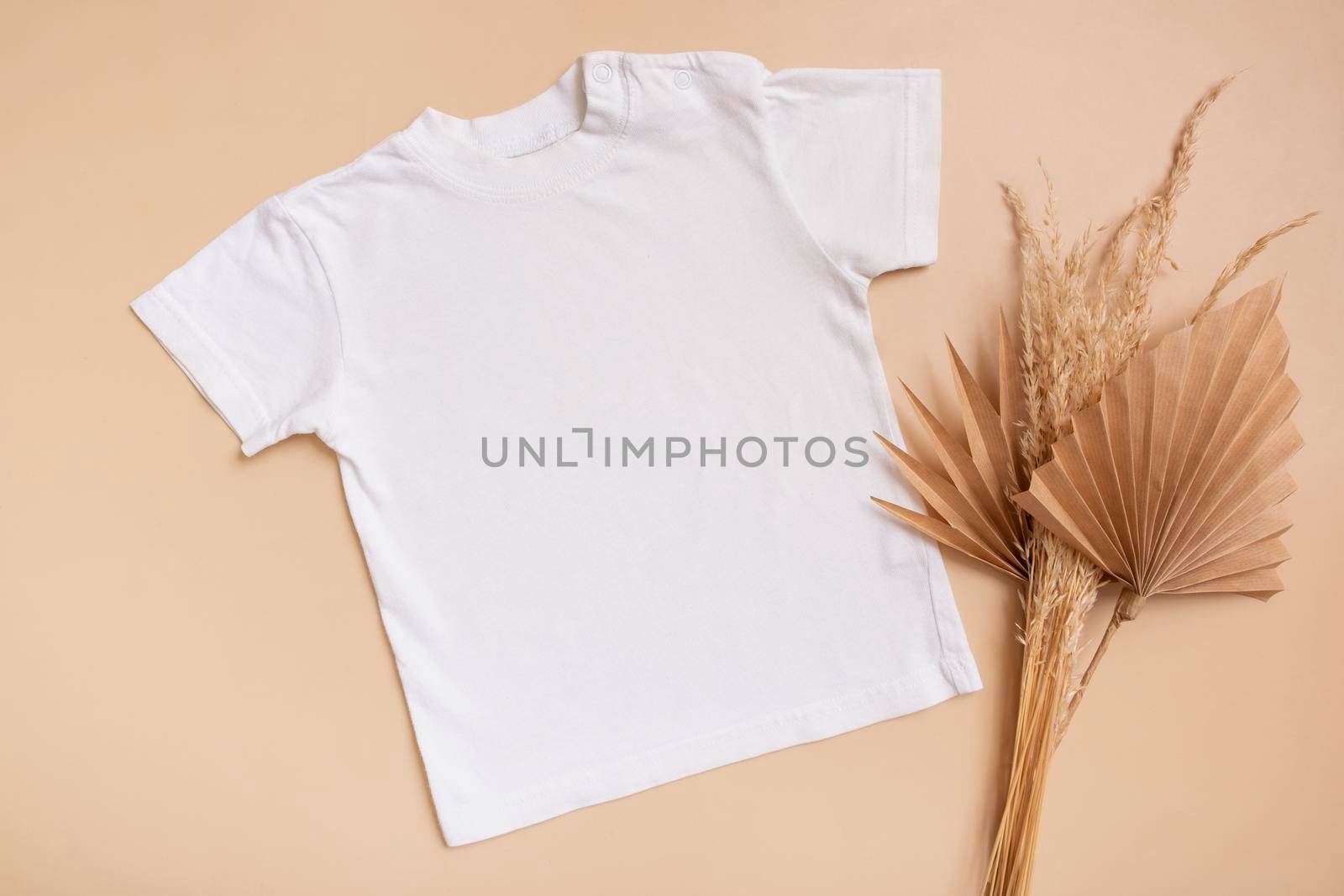 White baby t-shirt top view. Mock-up for logo or design on beige background. Flat lay child clothes with palm leaves by ssvimaliss