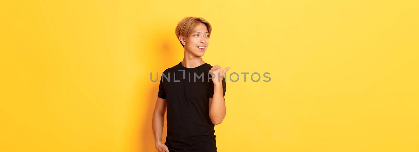 Portrait of pleased thoughtful asian guy, looking and pointing finger left at logo, standing yellow background by Benzoix